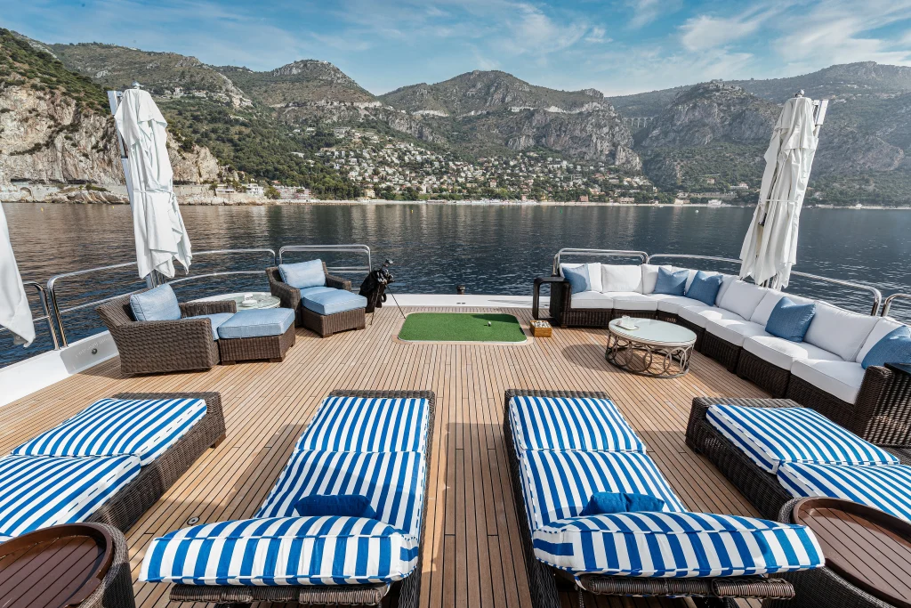 Four sun beds on deck with blue and white striped cushions. A long yacht sofa and two seats with cushions on the exterior deck. Two white sail awnings are tied up, and a small golfing green looks out over the water.