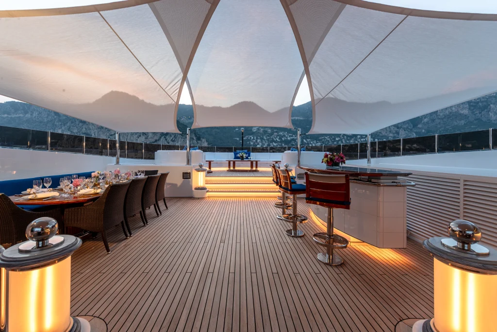 Large sail awning over the deck of a superyacht. Under is a bar with some stools and table and chairs.