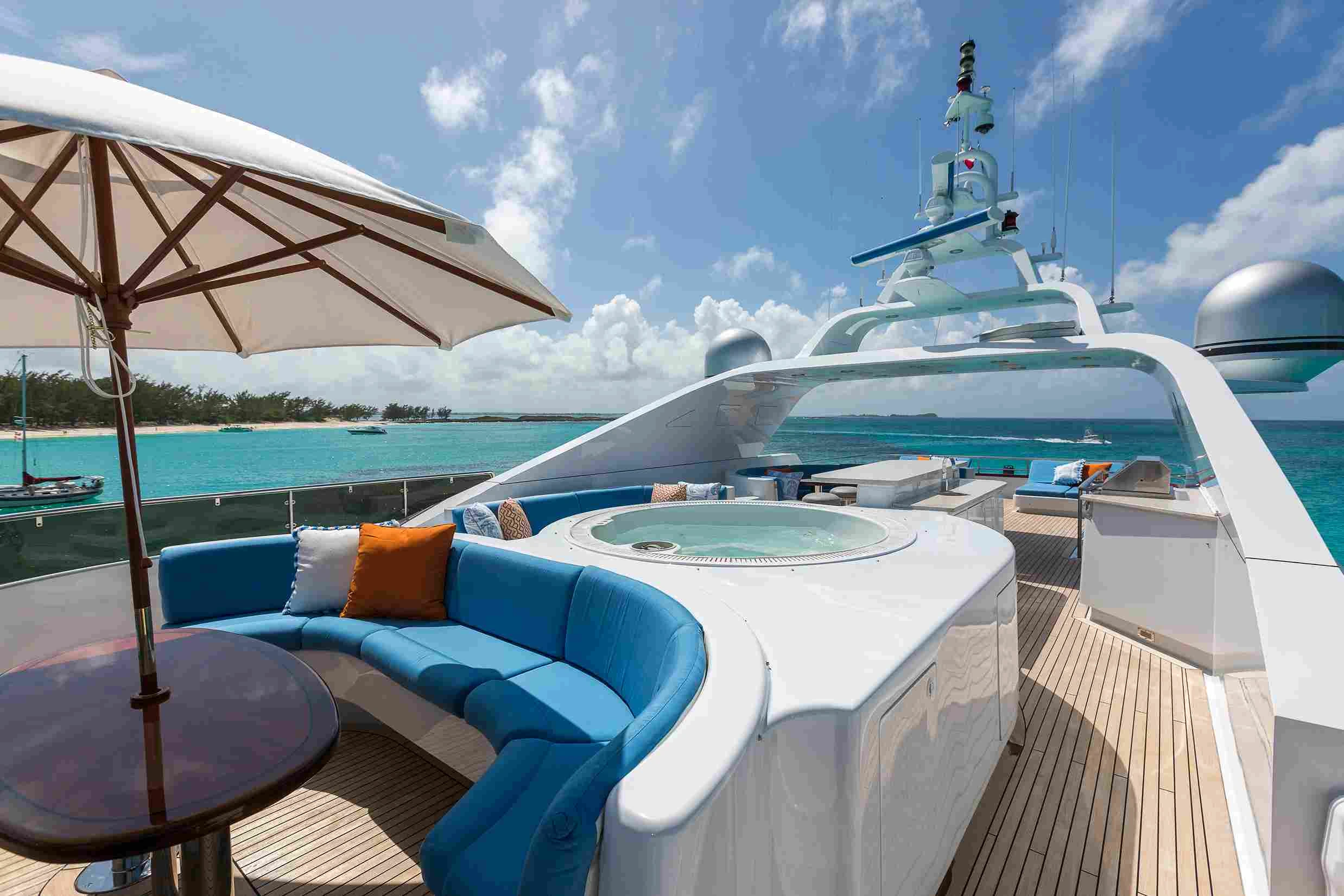 Blue yacht cushioned seating in front of a jacuzzi on a superyacht. It is a bright sunny day and a small sun awning is also open.